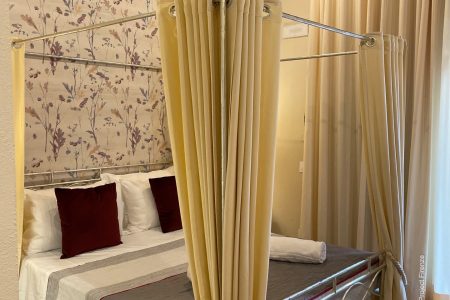 Un'accogliente camera da letto al Borgo Dolciano è dotata di un letto a baldacchino con tende gialle e cuscini bordeaux. La parete dietro il letto vanta una carta da parati floreale, che esalta il fascino rustico di questa Suite Mustiola con travi in legno a vista e un muro di mattoni parzialmente visibile.