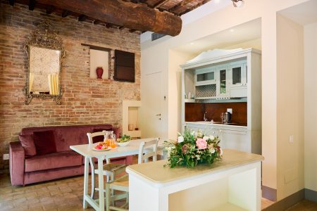 Un'accogliente cucina e zona giorno all'Agriturismo Chiusi presenta una parete in mattoni a vista, uno specchio vintage e un divano in velluto. Un tavolo da pranzo con sedie bianche è decorato con fiori. La cucina ha mobili bianchi e un piano di lavoro in legno con una composizione floreale.