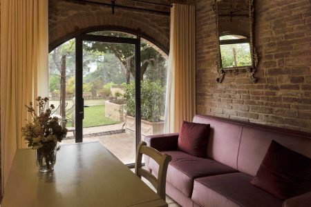 Accogliente camera con un muro di mattoni e una grande finestra ad arco e una porta che conduce a un giardino, che ricorda un soggiorno in un agriturismo in Toscana. Un divano viola è accanto alla finestra e un vaso di fiori è sul tavolo di legno. Le tende leggere incorniciano magnificamente la vista serena del giardino esterno.